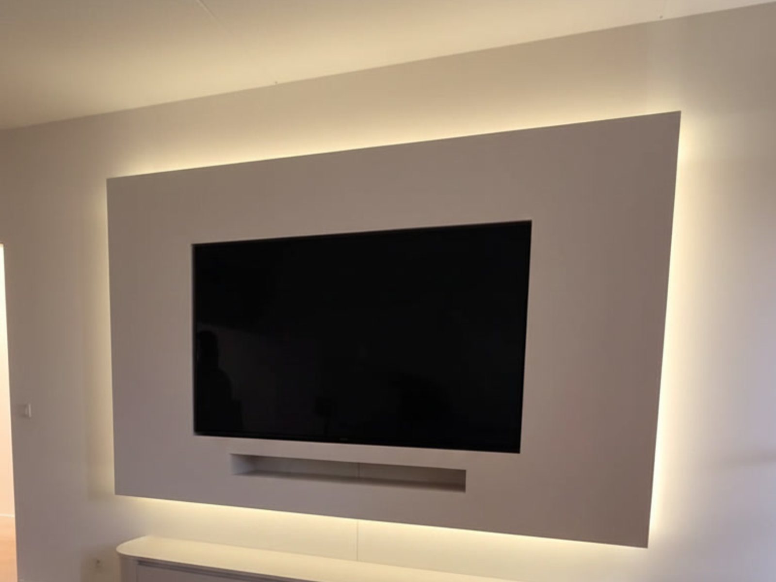 Cinewall zonder haard: strak wit wandpaneel met indirecte ledverlichting eromheen, een verzonken tv, soundbar-nis en een zwevend kastmeubel